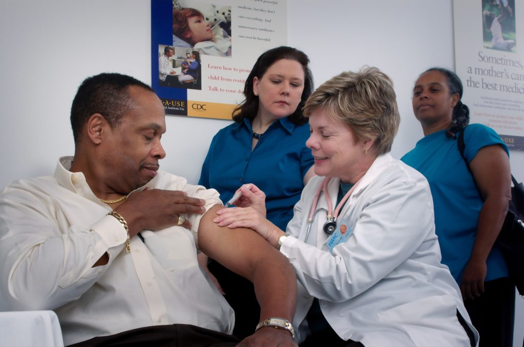 A healthcare professional administers a vaccination to a man, while others observe in a clinical setting.