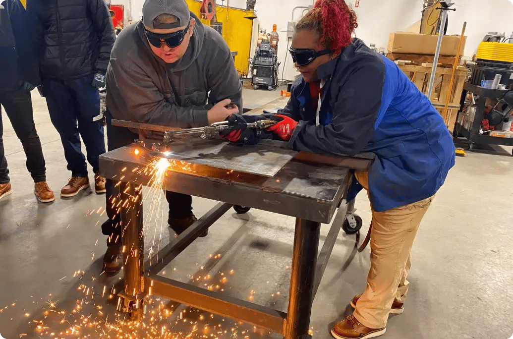 A woman in protective gloves uses a tool to cut metal, creating sparks, while colleagues observe in a workshop setting.