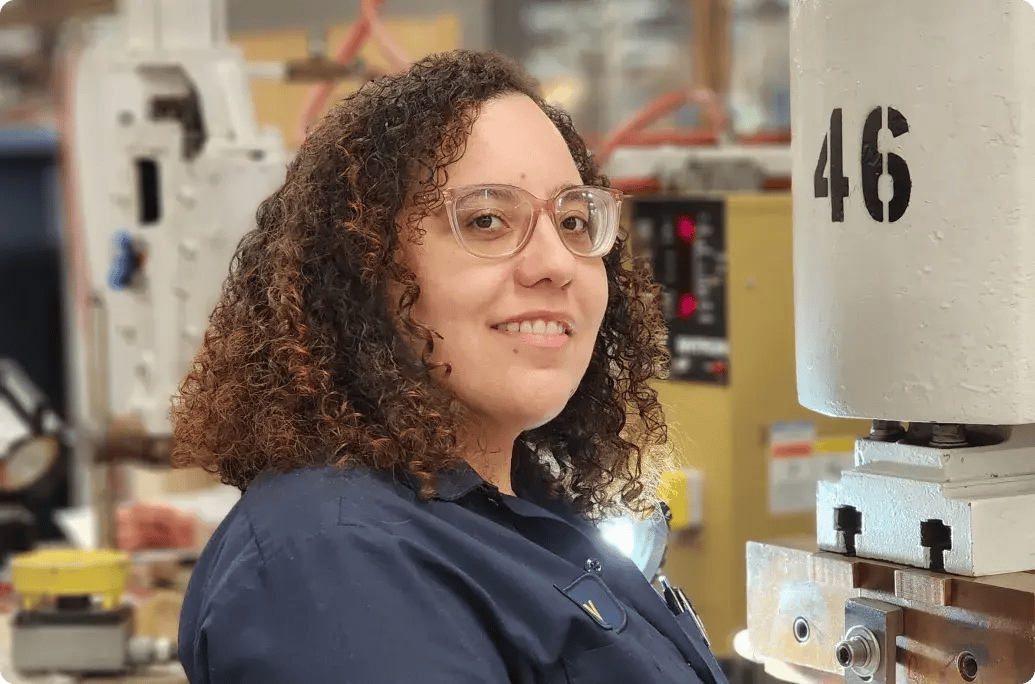 A person with curly hair wearing a blue shirt stands near industrial machinery, focused on their work environment.