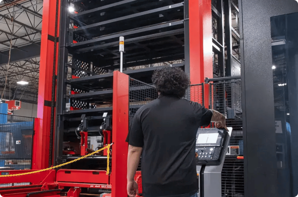 A worker operates a tall automated storage system with a control panel in a warehouse, surrounded by safety barriers.