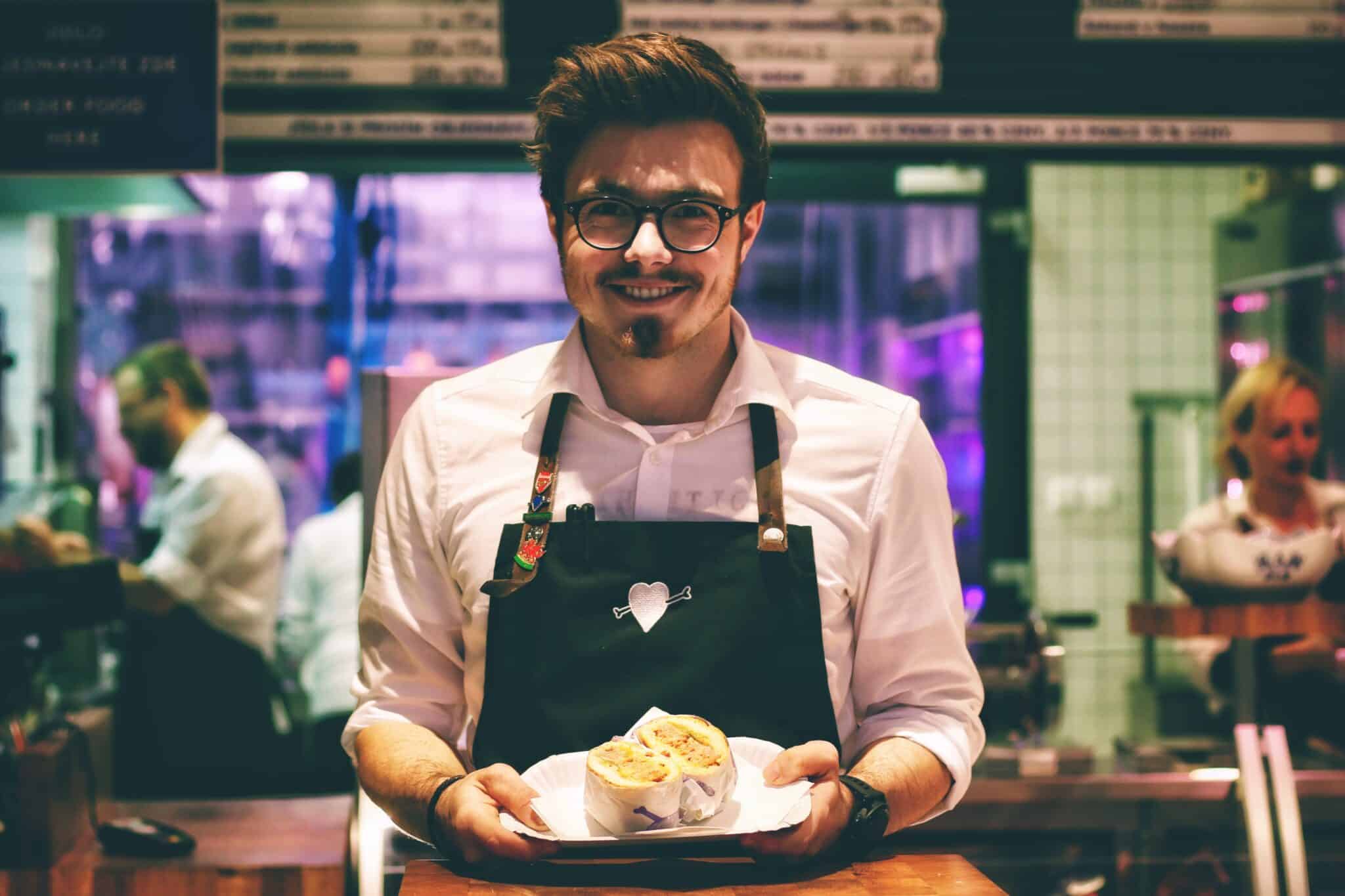 A staff member in an apron holds a plate with two wrapped food items in a vibrant, busy restaurant setting.