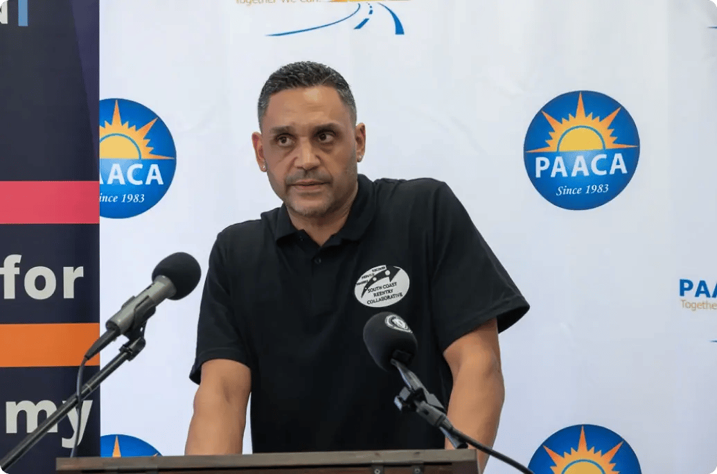 A speaker stands at a podium with microphones, addressing an audience against a backdrop featuring logos and a colorful banner.
