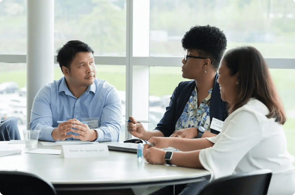 A diverse group of people engaged in a discussion around a table, with papers and cups, in a well-lit meeting room.