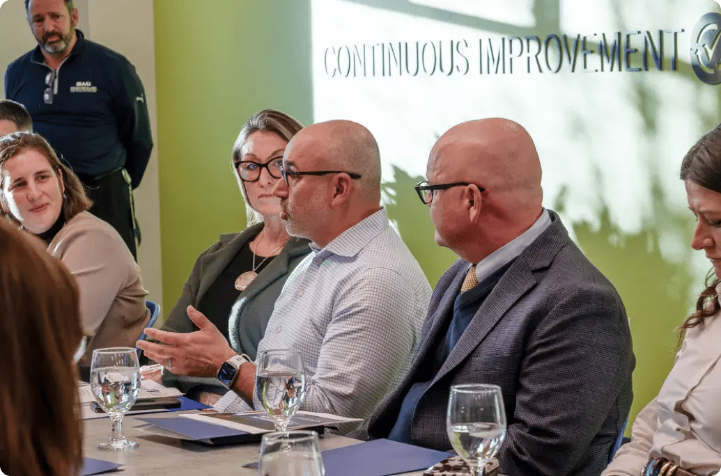 A group of professionals engaged in discussion around a conference table with water glasses and notebooks, focused on improvement strategies.