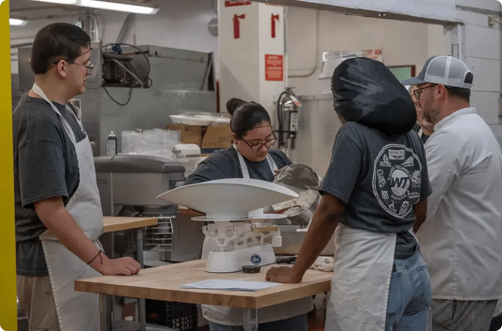 A group of people in a kitchen setting preparing ingredients, using a scale for measurements, surrounded by cooking equipment.