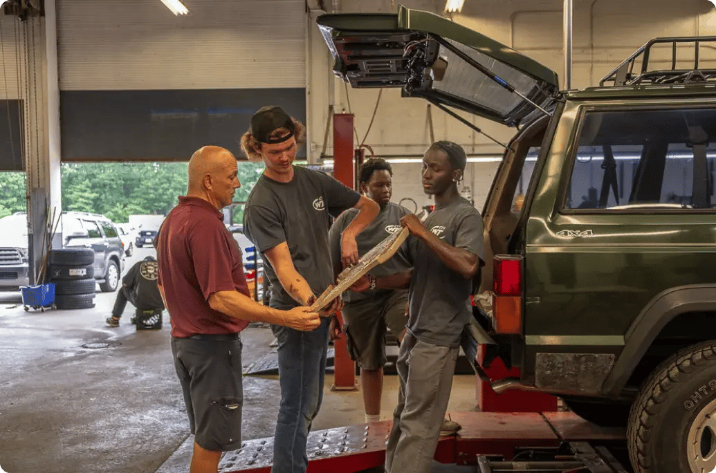 Four individuals are discussing a car part near an open hatchback of a green SUV in an auto repair shop.