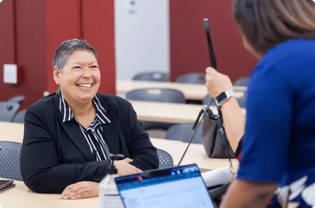 A woman in a black blazer sits at a table, while another woman in a blue shirt holds up a phone, talking in a classroom setting.