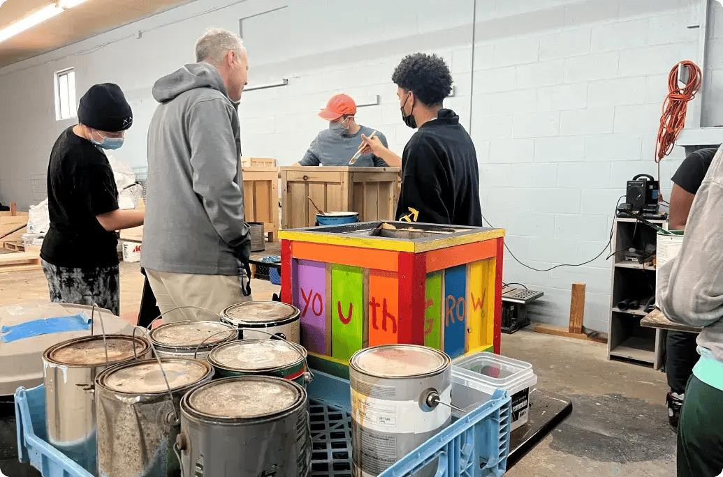 Participants work together in a workshop, painting colorful wooden boxes and discussing their project amidst paint cans and tools.